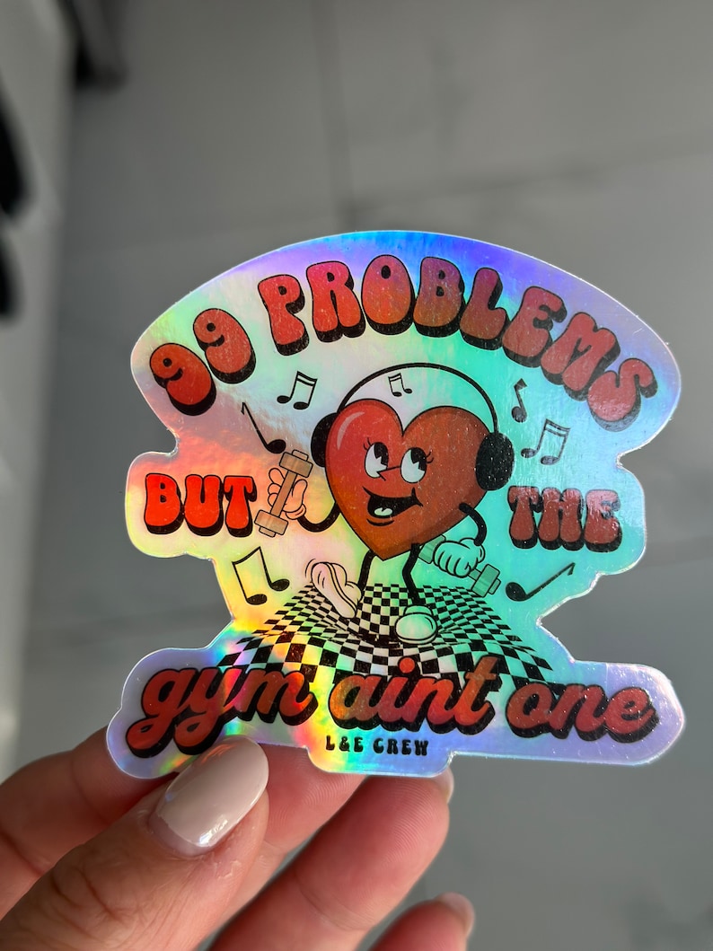 Puede incluir: Una pegatina hologr&aacute;fica con un coraz&oacute;n rojo que lleva auriculares y sostiene pesas. La pegatina dice "99 problemas pero el gimnasio no es uno" y "L&E Crew" en la esquina inferior derecha.