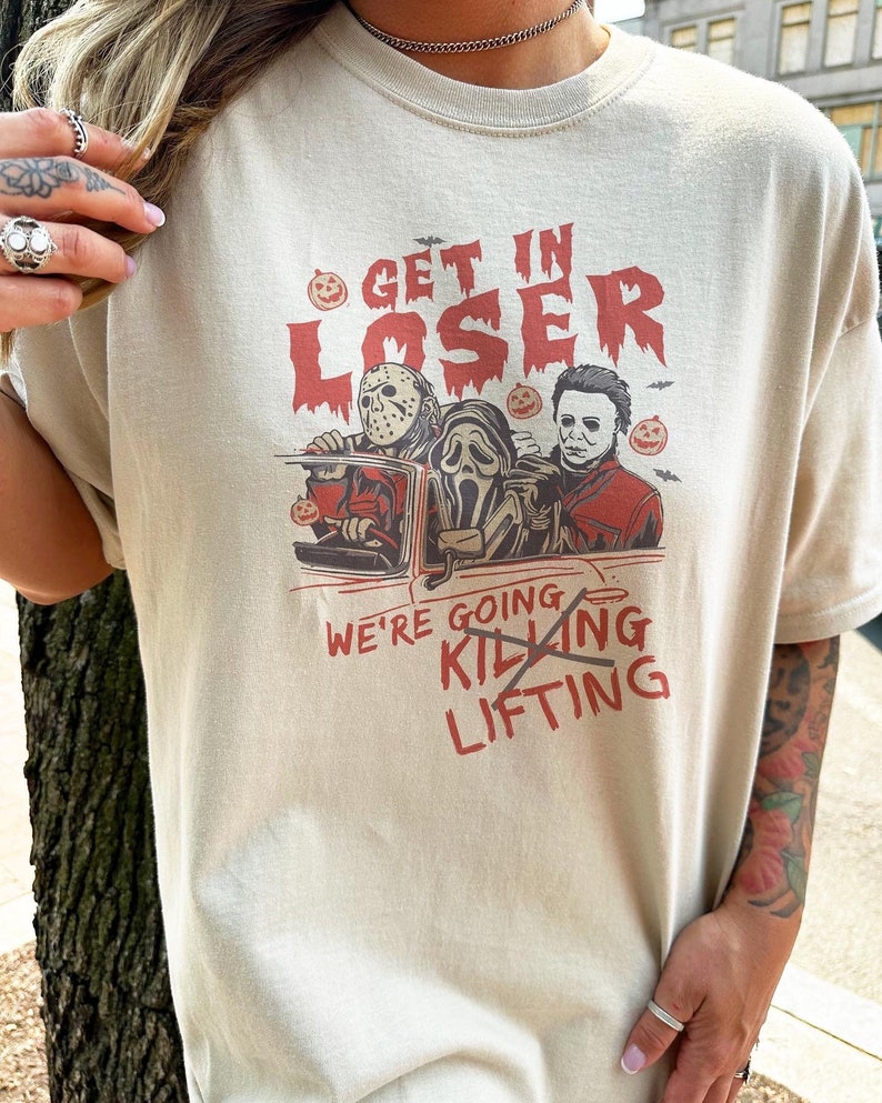 May include: A beige t-shirt featuring a graphic print of three horror movie characters in a car. The text "Get in Loser, We're Going Killing" is crossed out and replaced with "Lifting".