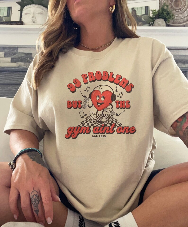 Puede incluir: Una camiseta beige con un gr&aacute;fico de un coraz&oacute;n rojo con auriculares y sosteniendo una mancuerna. El texto en la camiseta dice "99 problemas pero el gimnasio no es uno" con las palabras "LAE CREW" debajo.