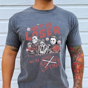 May include: A dark grey t-shirt featuring a graphic of three horror movie characters in a car. The text "Get in Loser" is above the characters and "We're going killing lifting" is below the characters. The text "killing" is crossed out.