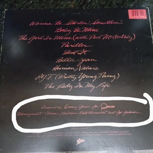 May include: Black and white album cover with the title "Thriller" by Michael Jackson. The album cover features a red handwritten list of songs including "Wanna Be Startin' Somethin'," "Baby Be Mine," "The Girl Is Mine (with Paul McCartney)," "Thriller," "Beat It," "Billie Jean," "Human Nature," "P.Y.T. (Pretty Young Thing)," and "The Lady In My Life." The album cover also includes the text "Produced by Quincy Jones for Qwest Records" and "Management: Meiner/Deben Entertainment and Joe Jackson."