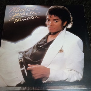 May include: A vinyl record cover featuring Michael Jackson in a white suit with a black shirt. The album title "Thriller" is written in gold lettering.