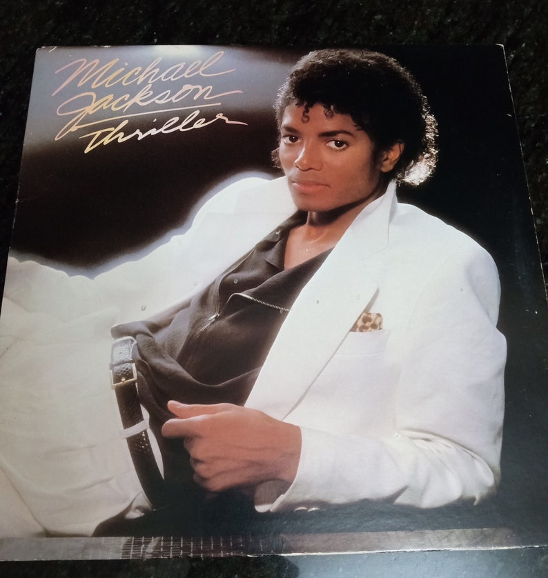 May include: A vinyl record cover featuring Michael Jackson in a white suit with a black shirt. The album title "Thriller" is written in gold lettering.