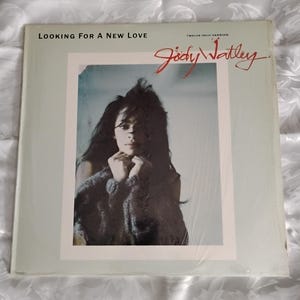 May include: A square vinyl record sleeve featuring a portrait of Jody Watley with dark hair, wearing a textured sweater. The text reads Looking For A New Love, Twelve Inch Version, and Jody Watley in red script against a light gray background.