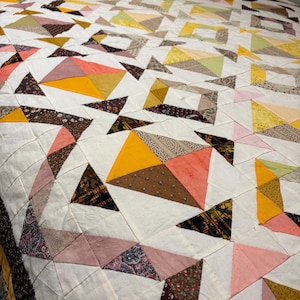May include: A patchwork quilt featuring a geometric pattern of triangles and squares. The design incorporates a variety of fabrics in shades of mustard yellow, coral pink, olive green, and dark brown, all set against a white background.