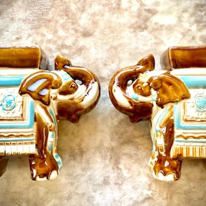 May include: A pair of ceramic elephant ornaments, each with brown, blue, and white detailing. The elephants feature decorative saddles and a seat on their backs. The ornaments are displayed together, highlighting their intricate design and craftsmanship.