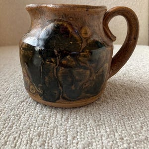 May include: Handmade ceramic mug with a brown and black mottled glaze. The mug has a rounded body, a curved handle, and a slightly flared rim. The glaze features dark, organic patterns.