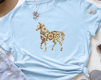 May include: Light blue t-shirt featuring a detailed horse graphic. The horse design incorporates various geometric patterns in shades of brown, beige, and green. The shirt is made of soft material and is suitable for casual wear.