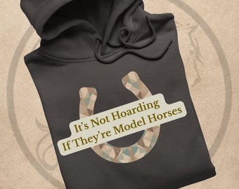 May include: Dark gray hoodie with a drawstring hood. The front features a horseshoe design with a geometric pattern in shades of brown, beige, and teal. Text on the hoodie reads "It's Not Hoarding If They're Model Horses."