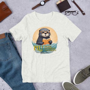 May include: A light grey t-shirt with a graphic of a cartoon otter wearing glasses and reading a book. The otter is floating in water with the text "PLU PACIFIC LUTHERAN UNIVERSITY MFA" below it.