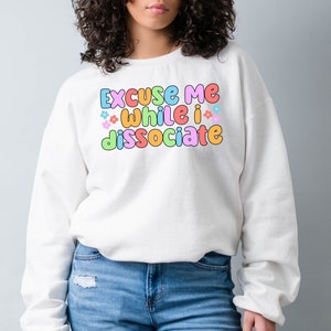May include: White sweatshirt with the text "Excuse me while I dissociate" in colourful bubble letters with small flower accents.