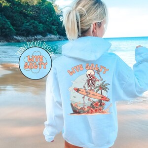 May include: A light blue hoodie with a graphic of a skeleton surfing on a surfboard with the text "Live Salty" on the back. The hoodie has a front pocket with the text "Live Salty" in a circle.