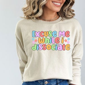 May include: A beige sweatshirt with the text "Excuse me while I dissociate" in colourful, outlined letters with small flower accents.