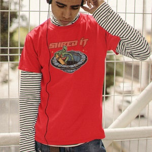 Pode incluir: Uma camiseta vermelha com um gr&aacute;fico de um alien&iacute;gena verde fazendo skate em um disco voador. O texto "Sliced it" est&aacute; impresso na camiseta.