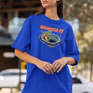 Pode incluir: Uma camiseta azul royal com um gr&aacute;fico de um alien&iacute;gena verde fazendo skate em um disco voador. O texto "Shred it" est&aacute; impresso em vermelho na camiseta.