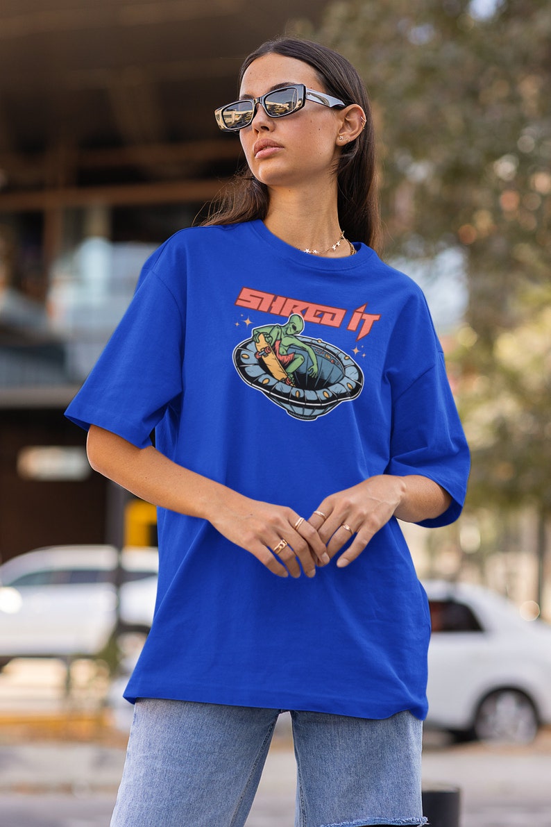 Pode incluir: Uma camiseta azul royal com um gr&aacute;fico de um alien&iacute;gena verde fazendo skate em um disco voador. O texto "Shred it" est&aacute; impresso em vermelho na camiseta.