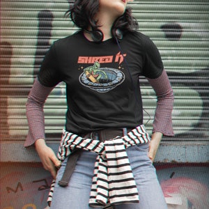 Pode incluir: Uma camiseta preta com um gr&aacute;fico de um alien&iacute;gena verde fazendo skate em um OVNI. O texto "Shred It" est&aacute; impresso acima do gr&aacute;fico.