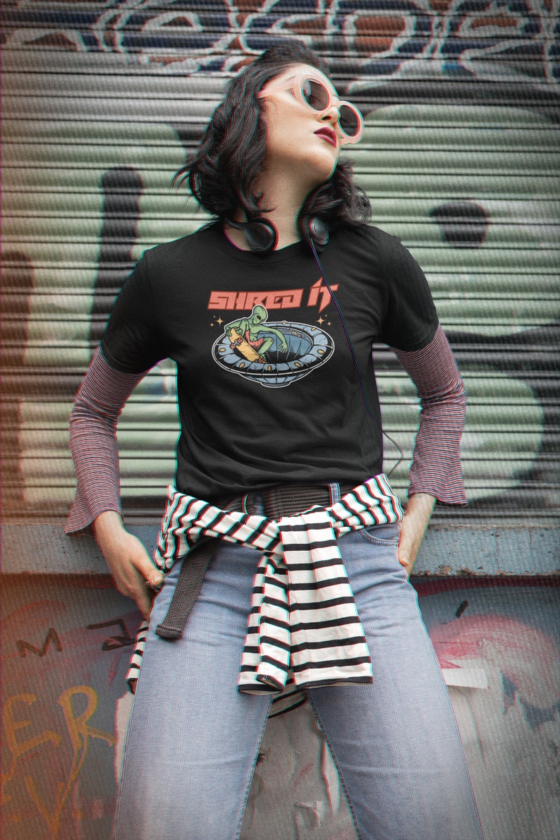 Pode incluir: Uma camiseta preta com um gr&aacute;fico de um alien&iacute;gena verde fazendo skate em um OVNI. O texto "Shred It" est&aacute; impresso acima do gr&aacute;fico.
