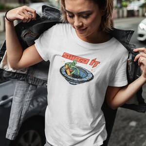 Pode incluir: Camiseta branca com um gr&aacute;fico de um alien&iacute;gena verde fazendo skate em um disco voador. O texto "Skate It" est&aacute; impresso em vermelho na camiseta.