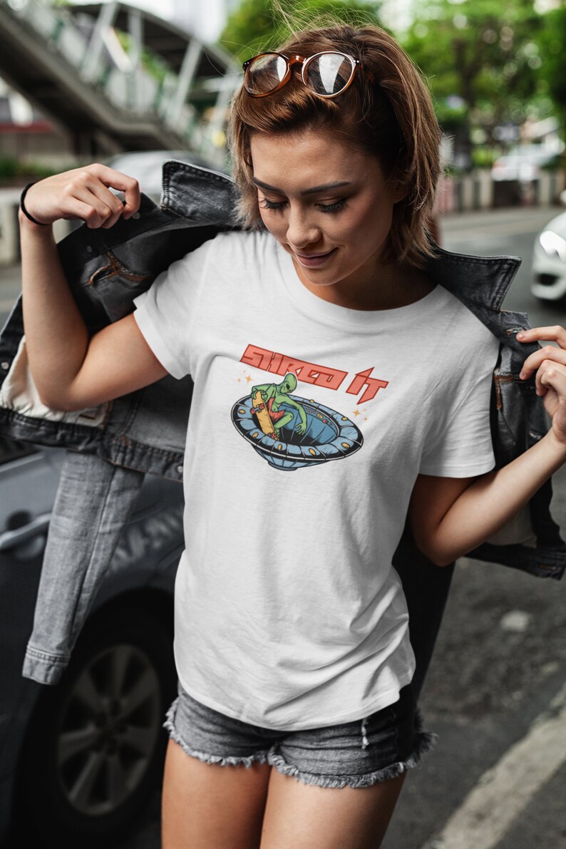 Pode incluir: Camiseta branca com um gr&aacute;fico de um alien&iacute;gena verde fazendo skate em um disco voador. O texto "Skate It" est&aacute; impresso em vermelho na camiseta.