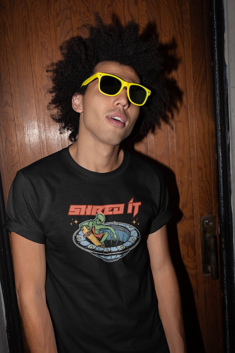 Pode incluir: Uma camiseta preta com um gr&aacute;fico de um alien&iacute;gena verde fazendo skate dentro de um disco voador. O texto "Shred It" est&aacute; impresso em vermelho acima do gr&aacute;fico.