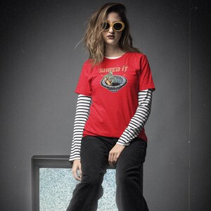 Pode incluir: Uma mulher vestindo uma camiseta vermelha com um gr&aacute;fico de um alien&iacute;gena verde em um disco voador e o texto "Shred It". Ela tamb&eacute;m est&aacute; vestindo uma camisa de manga comprida listrada de preto e branco por baixo e cal&ccedil;as pretas. Ela est&aacute; sentada em uma televis&atilde;o vintage com est&aacute;tica na tela.