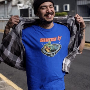 Pode incluir: Uma camiseta azul com um gr&aacute;fico de um alien&iacute;gena fazendo skate em um OVNI. O texto "Shred It" est&aacute; impresso em vermelho acima do gr&aacute;fico.