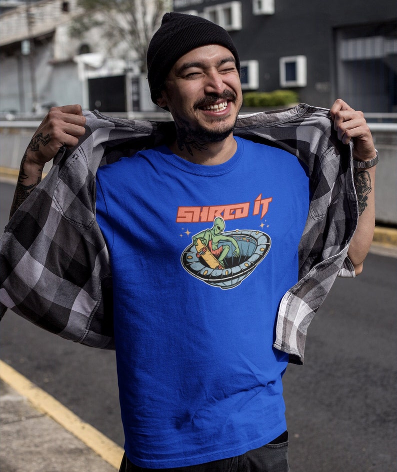Pode incluir: Uma camiseta azul com um gr&aacute;fico de um alien&iacute;gena fazendo skate em um OVNI. O texto "Shred It" est&aacute; impresso em vermelho acima do gr&aacute;fico.