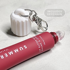 May include: A pink lip balm tube with the word "SUMMER" printed on it. The tube has a clear applicator tip and a silver keychain attached to the cap. The text "LIP BUTTER BALM BAUME A LEVRE Cherry | Cerise" is printed on the tube. The text "INCLUDES KEYCHAIN" is printed on a white background.