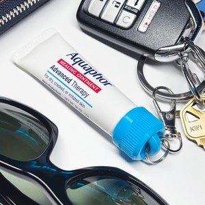 May include: A small blue and white tube of Aquaphor Healing Ointment Advanced Therapy, with the text "For dry, cracked, or irritated skin" and "Skin Protectant NET WT .35 OZ . 10g". The tube is attached to a silver key chain with a key and a car key fob.