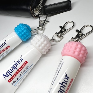 May include: Two Aquaphor Healing Ointment tubes with keychains attached. The tubes are white with blue, white, and pink caps. The text on the tubes reads "Aquaphor Healing Ointment Advanced Therapy".