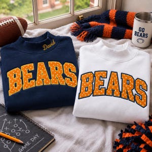 May include: A navy blue sweatshirt and a white sweatshirt, both featuring a mock neck and the word BEARS in orange with white polka dots. The navy sweatshirt has the name David embroidered in gold on the collar. A football, a tumbler with GO BEARS text, and a notebook are nearby.