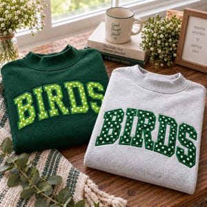 May include: A dark green sweatshirt and a light gray sweatshirt, both featuring a mock neck and the word BIRDS in large, block-style letters. The letters on the green sweatshirt are lime green with white polka dots, while the gray sweatshirt has dark green letters with white polka dots.