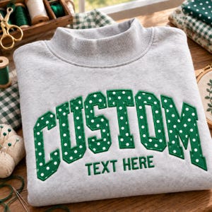 May include: A light grey sweatshirt featuring a mock neck collar and long sleeves. The front displays the word CUSTOM in large, green, white-polka-dotted varsity-style letters, with the words TEXT HERE embroidered in smaller green letters below.