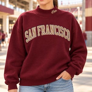 May include: A maroon long-sleeve sweatshirt with a mock neck collar. The text 49er is embroidered on the neck. The words SAN FRANCISCO are printed in a large, arched, gold-colored font with a white outline across the chest of the garment.