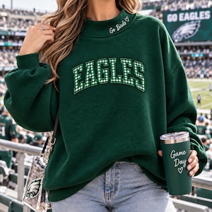 May include: A dark green crewneck sweatshirt featuring the word EAGLES in white and green gingham block lettering across the chest. The neckline includes the text Go Birds with a small heart. A matching dark green tumbler displays Game Day.