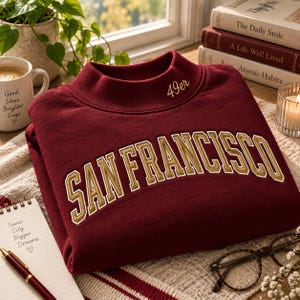 May include: A maroon crewneck sweatshirt featuring the text SAN FRANCISCO in large, gold-outlined block letters across the chest. The collar area includes the embroidered text 49er. The garment is made of a soft, solid-coloured fabric.