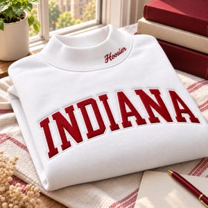 May include: A white mock neck sweatshirt featuring the word INDIANA in large, bold, red embroidered letters across the chest. The word Hoosier is embroidered in smaller red script on the collar. The garment has long sleeves and a ribbed neckline.
