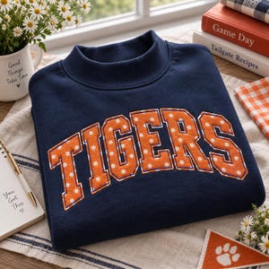 Pode incluir: Uma camisola azul-marinho de manga comprida com a palavra TIGERS em letras grandes de bloco laranja com bolinhas brancas e um contorno branco fino. A peça tem uma gola alta e é feita de um tecido de cor sólida.