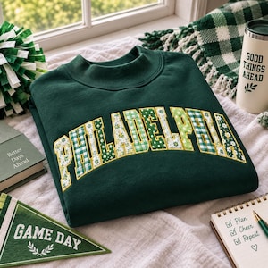 Peut inclure: Un sweat-shirt à col rond vert foncé avec le mot PHILADELPHIA écrit en grandes lettres majuscules arquées. Les lettres sont remplies d'un motif floral et vichy blanc, vert et jaune, souligné par une fine bordure jaune.