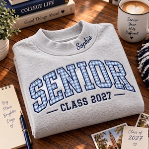May include: A light gray crewneck sweatshirt featuring the word SENIOR in blue and white gingham block letters, with the text CLASS 2027 underneath. The name Sophia is embroidered in dark blue script on the collar near the neckline.