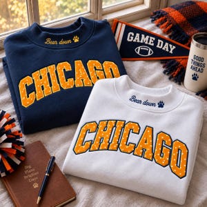 May include: A navy blue sweatshirt and a white sweatshirt, both featuring the word CHICAGO in orange with white polka dots. Each sweatshirt has the text Bear down and a paw print embroidered on the collar. A pennant reads GAME DAY.