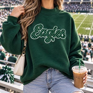 May include: A dark green long-sleeve sweatshirt featuring a mock neck and the word Eagles written in a white script font with a polka dot pattern. The sweatshirt is paired with light blue denim jeans and a small cream-colored shoulder bag.