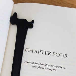 May include: A book with a black bookmark shaped like a dachshund dog. The book is open to Chapter Four, which reads "You can find kindness everywhere, even from strangers.". Clear glasses are resting on the book.