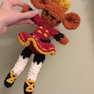 May include: A handmade crochet doll in the shape of a mouse. The doll has large orange ears, a red and yellow dress, and black and white legs with yellow accents. The doll is being held by a hand.