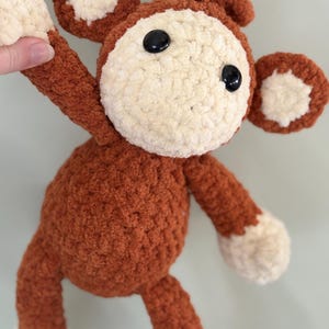 May include: A crocheted monkey toy with brown fur, white ears, and black eyes. The monkey is holding its arm up in the air.