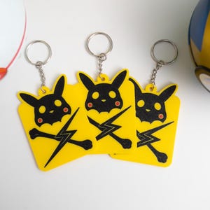 Może przedstawiać: Trzy żółte breloczki do kluczy z czarnym wzorem Pikachu i błyskawicą. Breloczki mają srebrne łańcuszki. W tle dwie ozdobne kule, jedna biało-czerwona, druga niebiesko-złota.