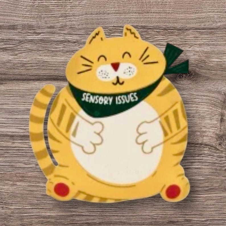 Pu&ograve; includere: Un gatto giallo con una bandana verde che dice "Sensory Issues". Il gatto sorride e ha una pancia bianca.