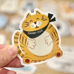 Pu&ograve; includere: Un gatto cartone animato giallo e arancione con strisce nere e una bandana nera che dice "Sensory Issues".