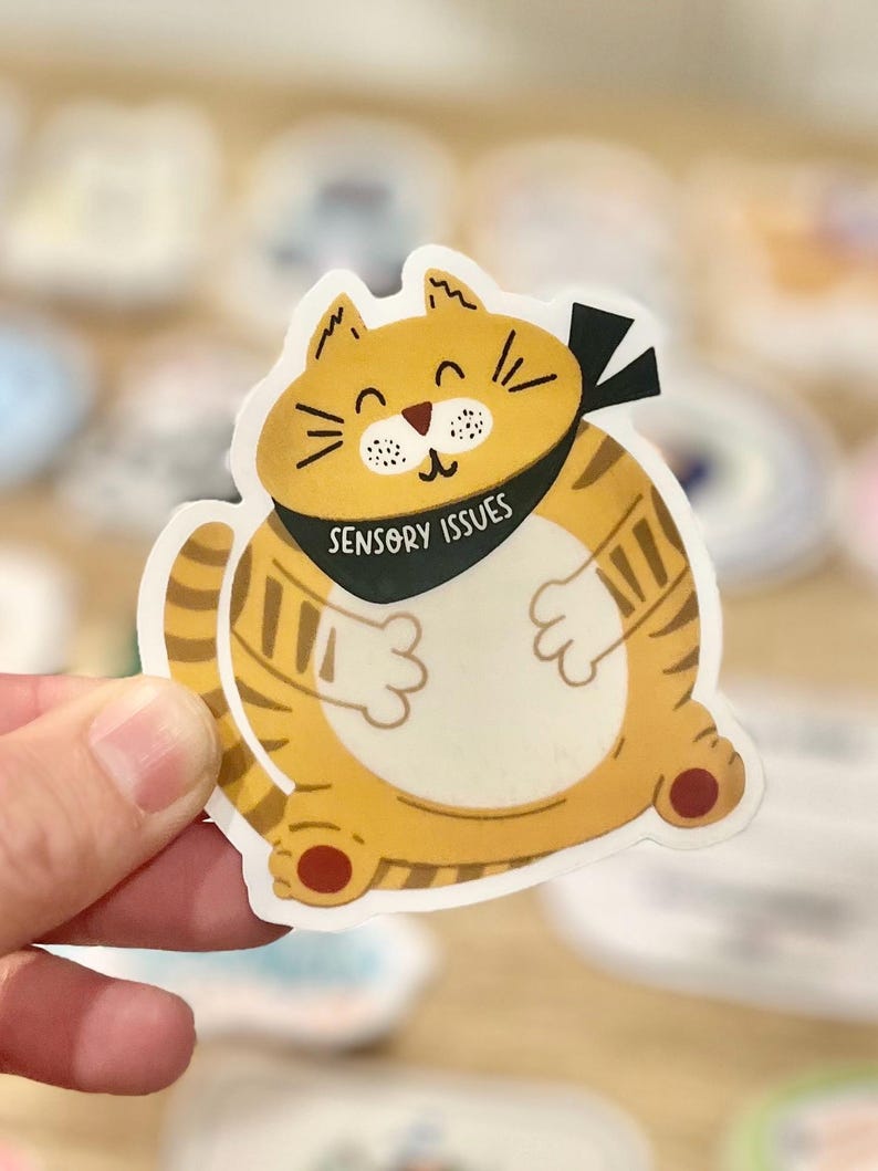 Pu&ograve; includere: Un gatto cartone animato giallo e arancione con strisce nere e una bandana nera che dice "Sensory Issues".